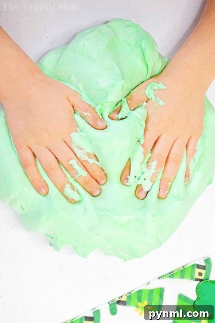 Explore the unique texture of shaving cream slime, a borax-free and kid-safe sensory activity for imaginative play. A child's hands gently pulling apart a mass of green fluffy shaving cream slime, showing its soft and airy texture.