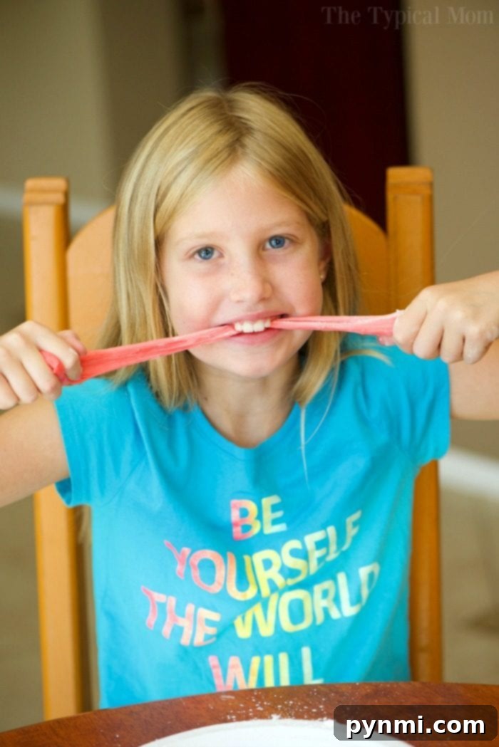 Cloud Slime with Shaving Cream 2 A child in a blue shirt stretches pink slime while sitting at a table, pondering how to make edible slime for an exciting twist.