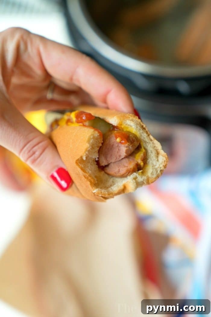 A hand holds a bitten hot dog with mustard and ketchup, the vibrant red nails adding a pop of color. Perfectly cooked using an Instant Pot, this hot dog promises both convenience and flavor in every bite.