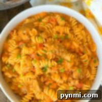 Hearty Instant Pot Goulash 2 A bowl of rotini pasta with creamy tomato sauce, bell peppers, and chives sits beside an Instant Pot and a floral napkin, reminiscent of a cheesy goulash twist.