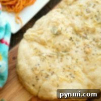 Homemade slow cooker rolls topped with herbs and shredded cheese sit on a wooden surface, while slow cooker rolls cool beside it. In the background, a corner of a white plate with spaghetti can be seen, complemented by a colorful cloth and a sprig of fresh herbs nearby.
