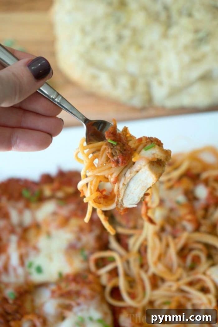 Close-up of a fork holding spaghetti and chicken over a plate of pasta, with freshly baked slow cooker rolls in the background.