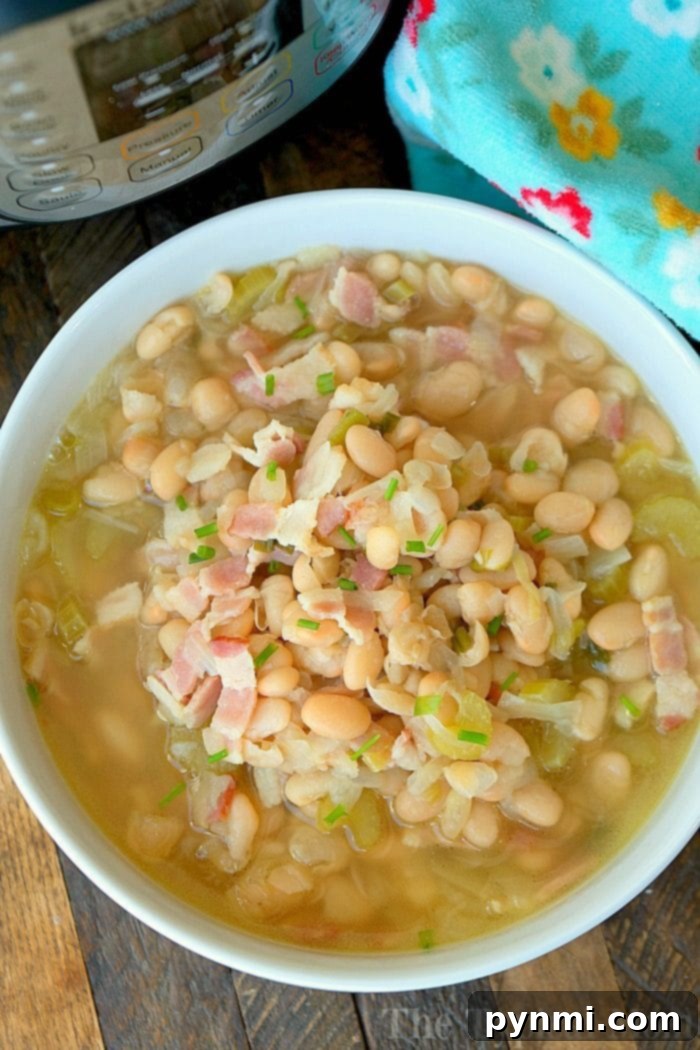 A steaming bowl of healthy pressure cooker bean soup, garnished with crispy bacon bits and fresh herbs, sits beside a decorative kitchen cloth.