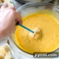 A bowl of yellow cheese fondue with broccoli invites a hand to dip a piece of bread using a blue fork. An Instant Pot pressure cooker sits in the background, while a plate with bread chunks waits on the side.