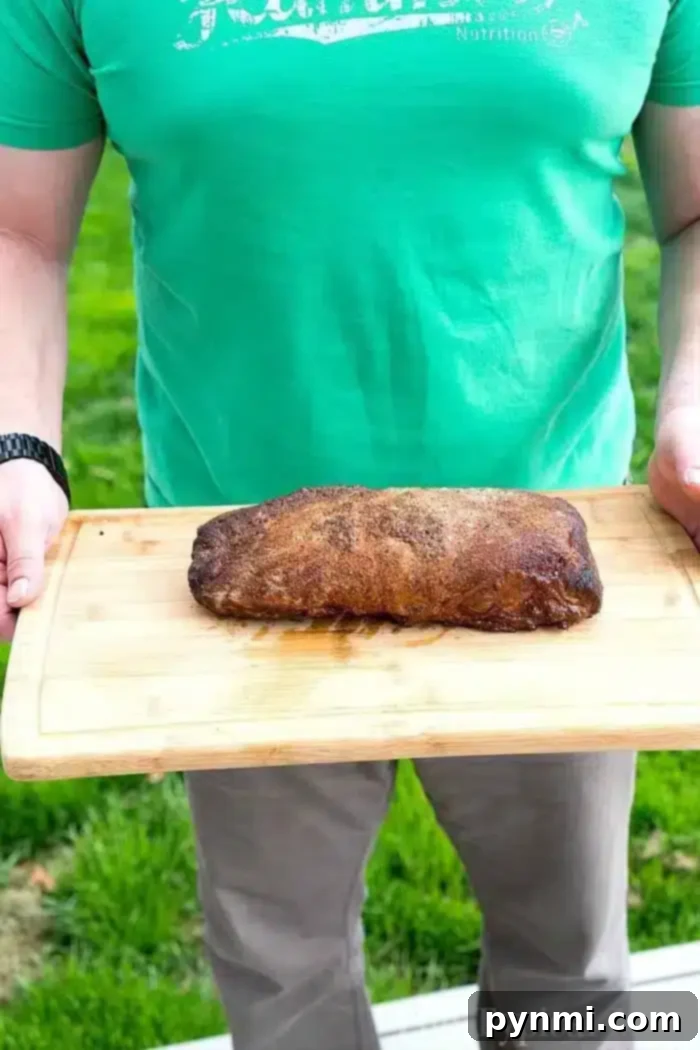 A person stands on the lush grass, holding a wooden board showcasing a perfectly smoked boneless pork loin.