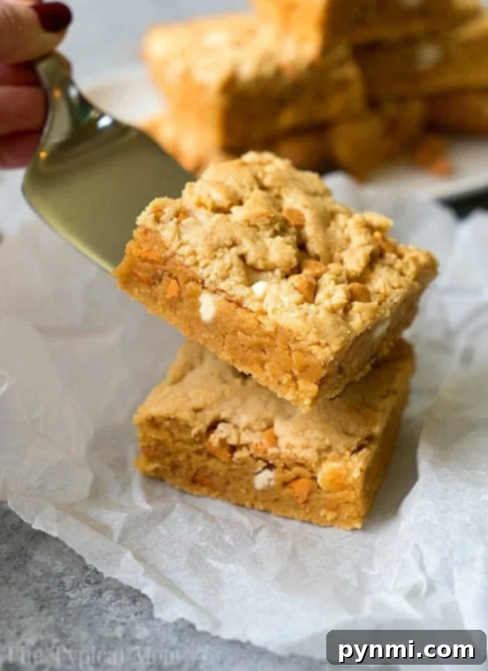 Ultimate Cake Mix Peanut Butter Brownies 2 A spatula lifts a square cake mix peanut butter brownie stuffed with peanut butter chips and white chocolate chips, placed on parchment paper, showcasing its rich, gooey texture and golden-brown edges.