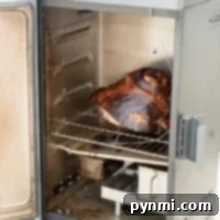 A metal smoker with the door open, revealing a large, perfectly smoked cooked ham on the rack inside, with visible smoke.