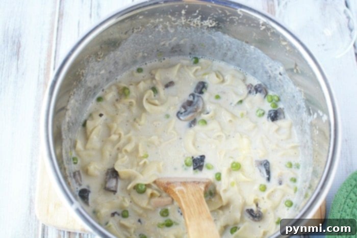 A pot of creamy tuna noodle pasta with mushrooms and peas, reminiscent of a classic pressure cooker tuna noodle casserole, is gently stirred with a wooden spoon on a rustic wooden surface.
