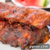 A plate with two racks of seasoned, grilled ribs showcases the best way to smoke ribs. In the background, there's a slice of watermelon on a table.