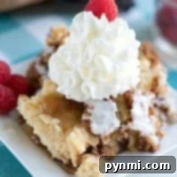 A delightful apple spice dump cake sits on the plate, topped with crumbled goodness, a dollop of whipped cream, and a single raspberry. The background features vibrant raspberries and a quaint bowl, setting the perfect scene for this autumn-inspired dessert.