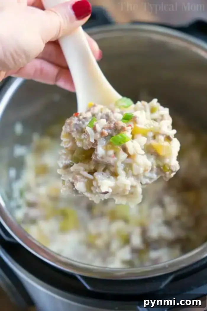 A hand holding a white spoon serves a portion of Instant Pot sausage rice casserole. The dish features creamy, seasoned rice blended with ground meat and green peppers, complemented by small vegetable pieces.