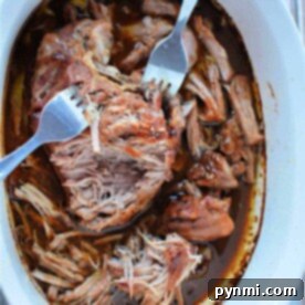 Succulent shredded pork in a white oval dish awaits, accompanied by two forks for easy serving. Master the art of cooking pulled pork and savor its tender goodness.