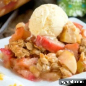 A delightful apple crisp topped with a scoop of vanilla ice cream rests on a white plate, accompanied by a floral napkin nearby. The subtle hint of rhubarb crunch adds an unexpected twist to this classic dessert experience.