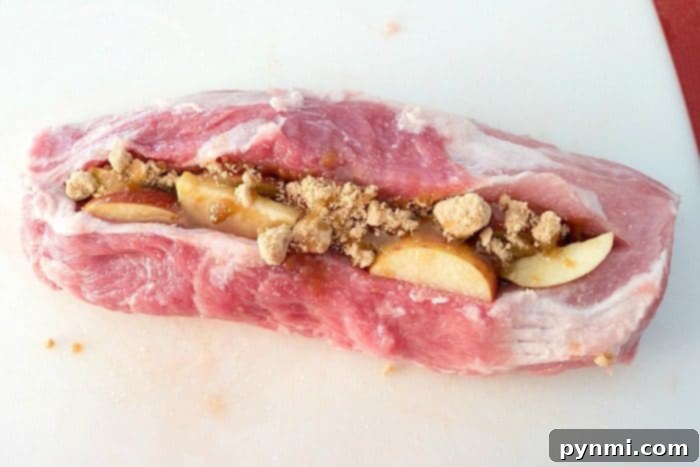 Succulent Smoked Stuffed Pork Loin 3 A raw center-cut pork loin, expertly butterflied and generously stuffed with thinly sliced apples and a sprinkle of brown sugar, rests on a pristine white cutting board, ready for a delicious smoked pork preparation.