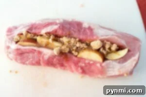 Raw pork loin, skillfully sliced open and generously filled with sweet apple slices and brown sugar, lies on a white cutting board—an essential step in preparing a mouthwatering smoked pork recipe.