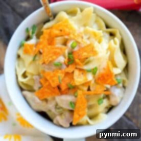 A delightful bowl of creamy pasta, a savory Dorito casserole, exquisitely garnished with finely crushed Doritos and freshly chopped chives. A spoon rests beside the bowl, accompanied by a bag of Doritos, hinting at the key ingredient.
