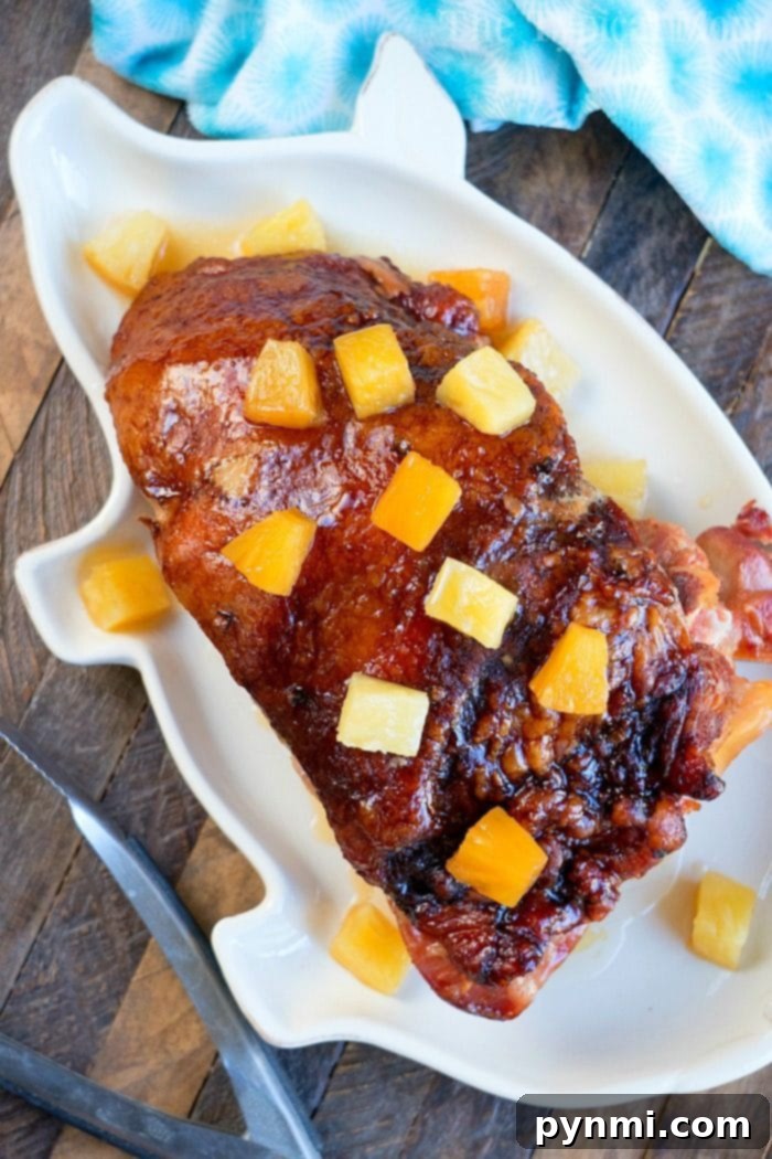 A beautifully glazed ham, adorned with golden pineapple chunks, resting on a pristine white platter, evoking the richness of a slow cooker ham recipe. The elegant display is set upon a rustic wooden table, complemented by a soft blue napkin.