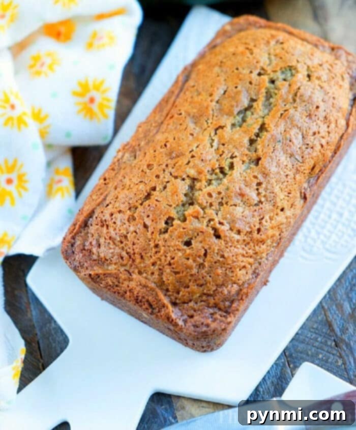 A perfectly golden-brown loaf of Grandma's moist zucchini bread on a white cutting board, beside a cheerful yellow floral napkin. This best zucchini bread recipe promises a delightful experience.