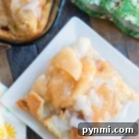 A plate of apple pie bread pudding with glaze rests beside a green floral napkin and a baking dish.
