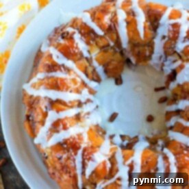 Gooey Cinnamon Apple Pull Apart Bread 3 A round pastry drizzled with white icing sits on a white plate, evoking the cozy delight of apple monkey bread made from cinnamon rolls. Juicy apples peek from the background, completing the scene.