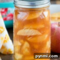 A jar of homemade pressure cooker apple pie filling rests on a wooden table, accompanied by a vibrant red apple and a delicate floral cloth, suggesting freshness and homemade goodness.