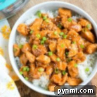 pressure cooker sesame chicken, perfectly glazed and sprinkled with green onions, rests atop a bed of rice on a wooden surface. A floral napkin adds an elegant touch to the setting.