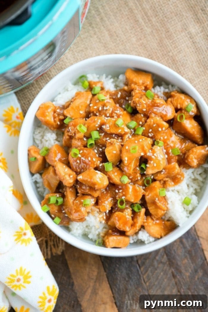 Pressure cooker sesame chicken, perfectly glazed and sprinkled with green onions, rests atop a bed of rice on a wooden surface. A floral napkin adds an elegant touch to the setting.