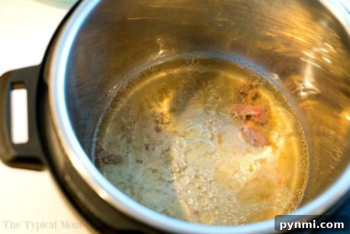 A block of frozen ground beef on a trivet inside an Instant Pot, surrounded by broth and seasonings for a flavorful cook.