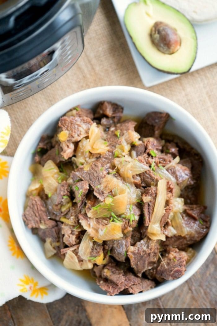 French onion pressure cooker roast is tender with tons of flavor. Perfect Instant Pot shredded beef tacos for dinner even your kids will rave about. #instantpotrecipes #instantpot #roast #frenchonion #tacos #shreddedbeef #ninjafoodirecipes #crockpotexpressrecipes #pressurecookerrecipes A bowl of shredded French onion beef with onions, ready to be served, accompanied by an avocado half on a rustic wooden surface.