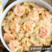 A bowl of creamy instant pot shrimp Alfredo with spiral-shaped noodles, garnished with grated cheese and small bits of herbs. The dish is placed on a wooden surface with a napkin partially visible to the side.