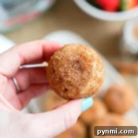 A hand holds a round, cinnamon-sugar-coated treat, reminiscent of pressure cooker churro bites. In the blurred background, more of these tasty delights sit alongside a bowl brimming with strawberries.