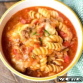 A bowl of rotini pasta, dubbed Ninja Foodi taco pasta, is topped with a chunky tomato and ground meat sauce, garnished with melted cheese and herbs. The bowl rests on a wooden surface beside a green cloth, with part of a kitchen appliance visible in the background.