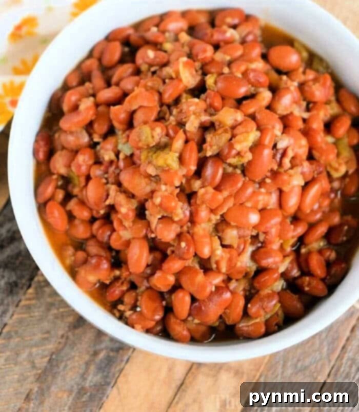 Rapid No Soak Pinto Beans Pressure Cooker 3 A white bowl filled with seasoned pinto beans, fresh from the pressure cooker, sits on a wooden surface. The reddish-brown beans are tantalizingly presented alongside a sliver of floral-patterned cloth peeking in from the top left corner.