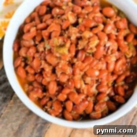 A white bowl filled with seasoned pinto beans, fresh from the pressure cooker, sits on a wooden surface. The reddish-brown beans are tantalizingly presented alongside a sliver of floral-patterned cloth peeking in from the top left corner.