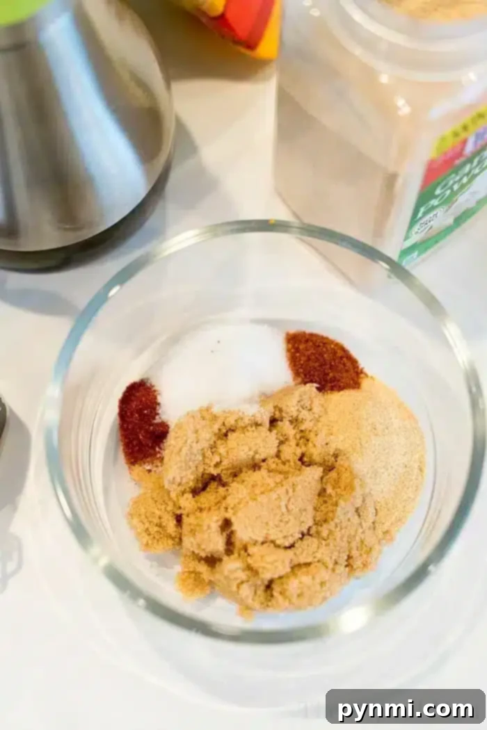 Best air fryer chicken thighs ever! Use this dry rub for chicken to make sweet and crispy chicken in an air fryer that's tender and juicy inside. Yum! #airfryerrecipes #airfryer #ninjafoodirecipes #ninjafoodi #chickenthighs #chicken #crispy A glass bowl containing various seasonings, including brown sugar, garlic powder, paprika, and salt. A metal object and a container of spices are partially visible in the background on the white surface—perfect for flavoring bone-in air fryer chicken thighs.