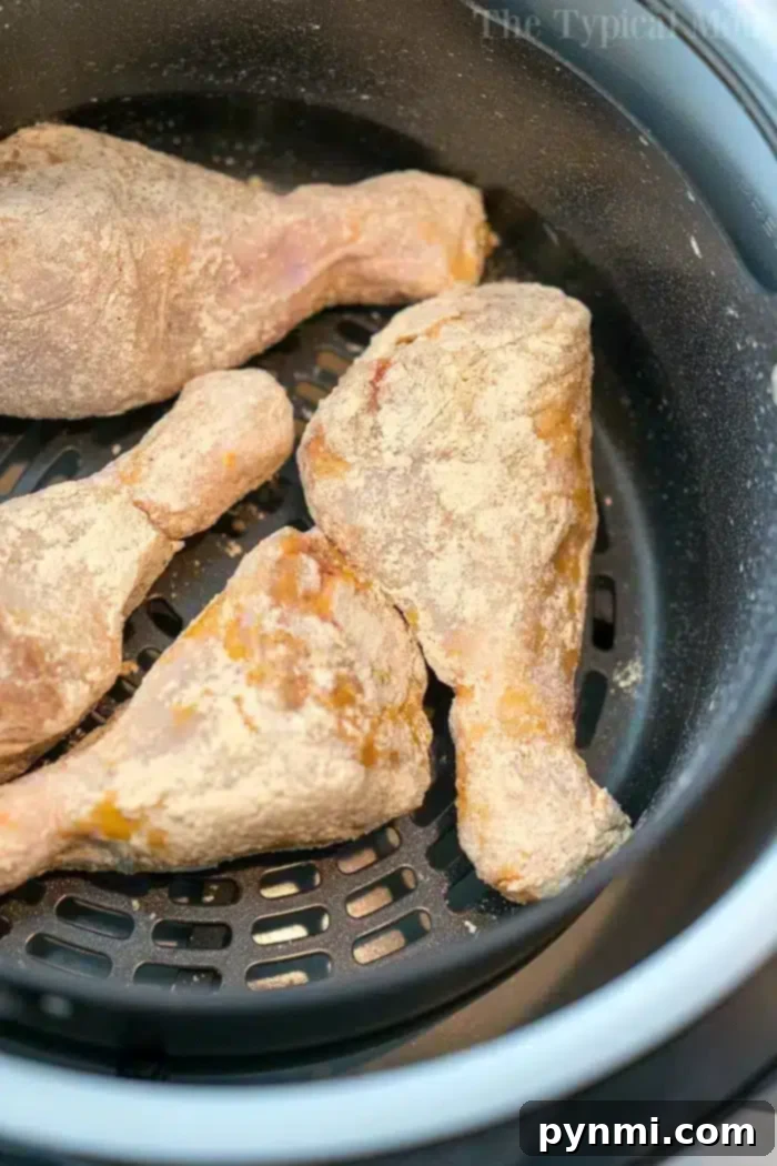 Chicken Drumsticks Ready for Air Frying Close-up of four raw, breaded chicken drumsticks carefully arranged in the black mesh basket of an air fryer, poised for cooking. The light coating of flour is visible, perfectly prepped for an easy air fryer fried chicken recipe. The setup highlights the simplicity and readiness of the meal.