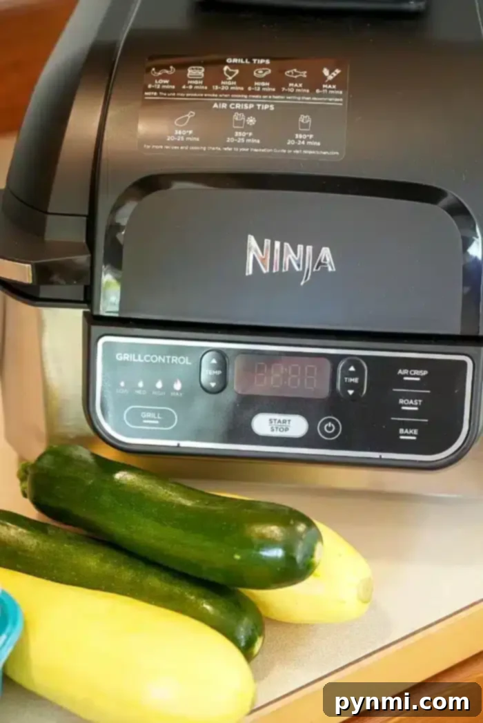 Best Ninja Foodi Grill vegetables you will ever make are here! No breading, just seasonings and mixed vegetables cooked to perfection. A healthy side dish fave. #ninjafoodi #airfryer #airfryerrecipes #vegetables #mixedvegetables #ninjafoodirecipes #ninjafoodigrill #mushrooms #zucchini #yellowsquash A Ninja air fryer is displayed on a kitchen counter, with two zucchinis and a yellow squash positioned in front of it. The appliance features a digital control panel and clear labels for grill and air crisp functions on its lid.