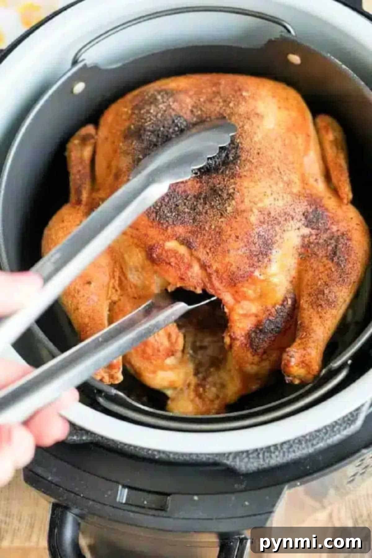 An air fryer whole chicken sits in the basket, perfectly cooked and golden, as metal tongs lift it up for serving.