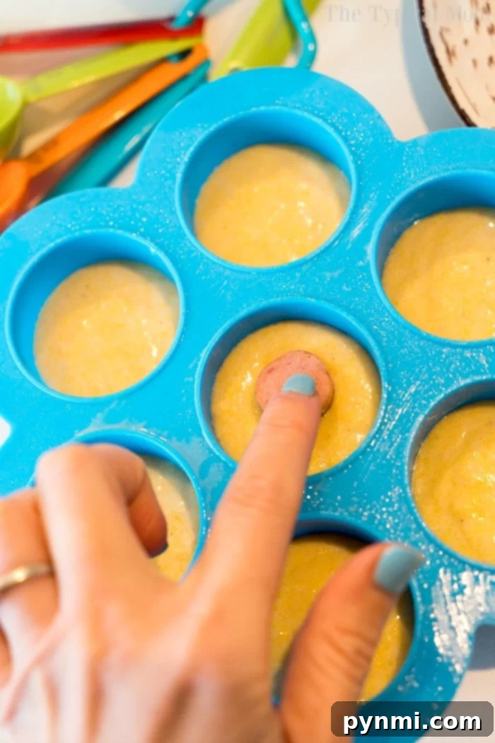 Pressure cooker corn dog bites are easy to make in your Instant Pot, Crock Pot Express or Ninja Foodi! The perfect snack or lunch your kids will love. #instantpotrecipes #instantpot #ninjafoodi #ninjafoodirecipes #corndogs #eggmold #pressurecooker #snacks #lunch #corndogbites A hand carefully places a mini hot dog piece into cornbread batter in a blue silicone egg mold, ready for pressure cooking.