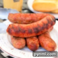 Smoked brats stacked on a white plate with tongs and buns in the background.