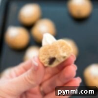 Close-up of a hand holding a half-eaten ball of edible raw cookie dough with a chocolate chip peeking out. Other dough balls and a baking tray are blurred in the background, tempting you with their gooey promise.