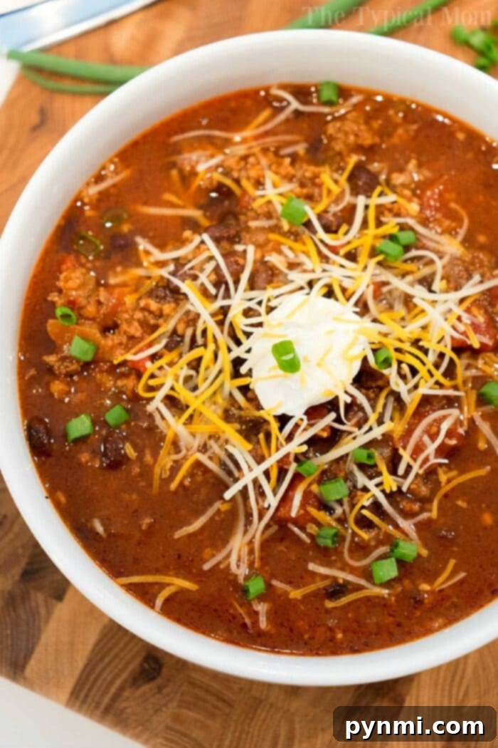 Bowl of Ninja Foodi chili topped with shredded cheese, sour cream, and chopped green onions on a wooden surface—a perfect mild chili recipe for cozy nights.