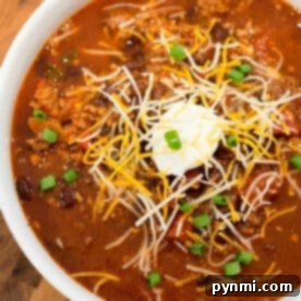 Bowl of Ninja Foodi chili topped with shredded cheese, sour cream, and chopped green onions on a wooden surface—a perfect mild chili recipe for cozy nights.