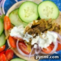 Plate with an instant pot chicken shawarma gyro sandwich, topped with sliced cucumbers, tomatoes, and onions, served alongside warm pita bread and a generous drizzle of sauce.