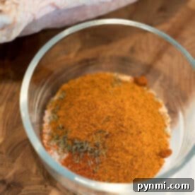 A glass bowl filled with a mix of dry spices for a smoked turkey rub recipe sits on a wooden surface next to raw poultry.