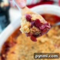 A hand holding a wooden spoon with a serving of tart cherry crisp over a white baking dish.
