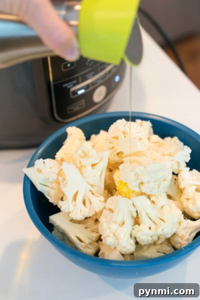 Easy air fryer cauliflower is easy to make in your Ninja Foodi or other machine. Healthy and full of flavor it's a great vegetable side dish or snack. #airfryer #ninjafoodi #cauliflower #vegetables #vegetarian #sidedish A stream of olive oil being drizzled over fresh cauliflower florets in a vibrant blue bowl, preparing them for air frying.