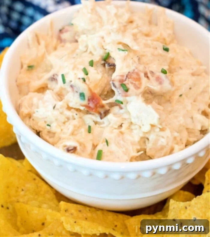 Ultimate Pressure Cooker Crack Chicken Dip 2 A white bowl brimming with creamy pressure cooker crack chicken dip, garnished with chopped chives. The bowl is surrounded by yellow tortilla chips on a table, inviting you to dive into this delicious creation.