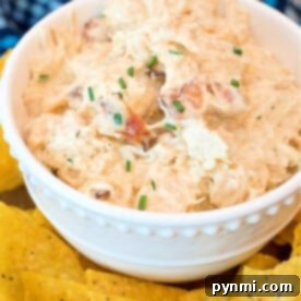A white bowl brimming with creamy pressure cooker crack chicken dip, garnished with chopped chives. The bowl is surrounded by yellow tortilla chips on a table, inviting you to dive into this delicious creation.