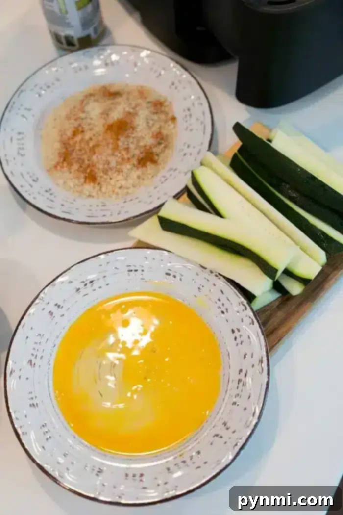 Two bowls on a white surface, one with beaten eggs, the other with a breadcrumb mixture. Next to the bowls are sliced zucchini on a cutting board, ready to become delicious air fryer zucchini fries.